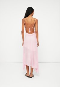 Ružová, priesvitná maxi šaty so špagetovými ramienkami a dizajnom so zaviazaním na chrbte. Má voľný siluetu a asymetrický lem. Nosené s plochými sandálmi.