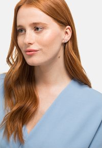 Roségoldene Ohrstecker mit einem gemusterten, kreisförmigen Design, getragen von einer Person mit langen, welligen roten Haaren und einem hellblauen Oberteil.