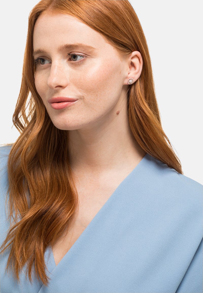 Roségoldene Ohrstecker mit einem gemusterten, kreisförmigen Design, getragen von einer Person mit langen, welligen roten Haaren und einem hellblauen Oberteil.