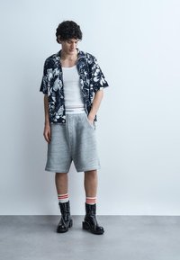 Mladi oseba oblečena v cvetlično srajco s kratkimi rokavi, brezrokavnik bele barve, sive kratke hlače, črtaste nogavice in črne gležnjarje, stoji v notranjosti.