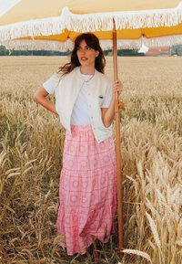 Un débardeur blanc sans manches sur un t-shirt blanc, associé à une jupe maxi à motifs roses avec des motifs de soleil, sous un parasol jaune à franges.