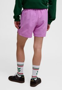 Lila elastiska shorts med sidopenningar och svarta zickzackmönster; ihop med vita strumpor med röda och gröna ränder samt mörka sneakers.