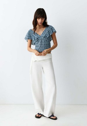 Femme portant un chemisier bleu à motifs épaules dénudées, un pantalon blanc taille haute à jambes larges, et des tongs noires, debout devant un fond blanc.