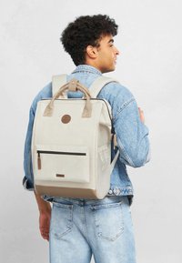 Mochila de lona beige con un asa superior, correas ajustables, un bolsillo frontal con cremallera y un acento en la parte inferior de cuero sintético. Detalle del logo redondo en la parte posterior.
