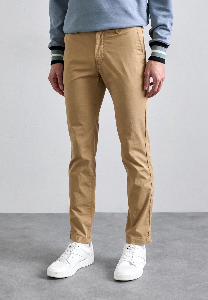 Khaki pants featuring a slim fit, made of smooth cotton fabric, with a front pocket and a tapered leg, paired with white sneakers.