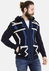 Marineblaue Strickjacke mit einem weißen und blauen geometrischen Muster, gerippten Bündchen und einem Reißverschluss. Hat große Taschen und strukturierte Ärmel.