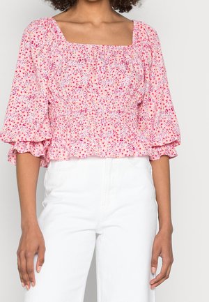 Rosa blütenmusterte Bluse aus weichem Stoff, mit quadratischem Ausschnitt, gerafftem Oberteil und Puffärmeln. Getragen mit weißen Hosen.