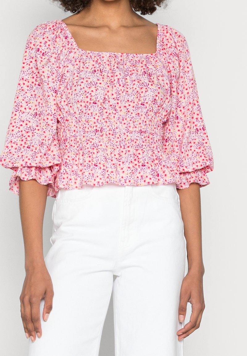 Blusa a fiori rosa, realizzata in tessuto morbido, con scollatura quadrata, corpetto arricciato e maniche a bufalo. Indossata con pantaloni bianchi.