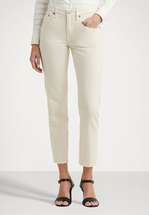 Jeans beige a vita alta con design aderente, stile a cinque tasche e chiusura a bottoni, abbinati a sandali neri con cinturini.