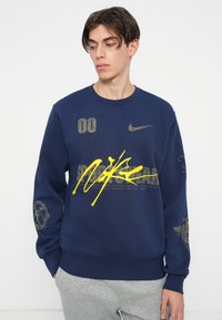 Mornarsko modra športna majica iz mešanice bombaža z zlatimi in rumenimi grafikami. Ima krožni izrez, dolge rokave in rebraste manšete. Dodatki vključujejo logotipe in številke.