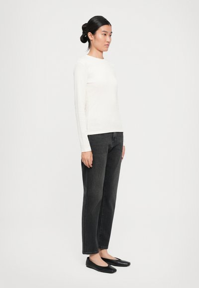 Kvinna som står i profil iförd en vit långärmad topp, svarta raka jeans och svarta lägenheter mot en enkel ljus bakgrund.