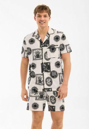 Młody mężczyzna w białej koszuli z krótkim rękawem i szortach z czarnymi tropikalnymi i słonecznymi wzorami, uśmiechający się na jednolitym tle.