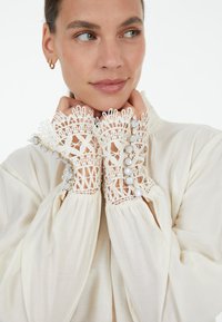 Lace-cuffed cream blouse with textured sleeves, featuring pearl-like buttons along the cuffs and intricate lace detailing.