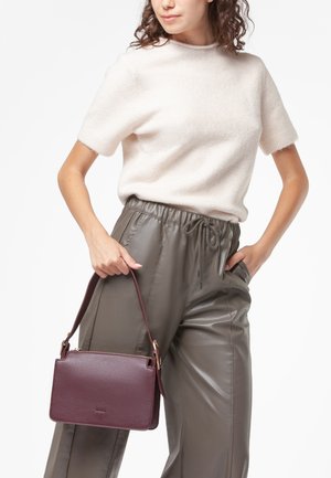 Borsa in pelle bordeaux con una forma strutturata, accessori in oro e una tracolla staccabile, tenuta da un modello che indossa un maglione chiaro e pantaloni in pelle.