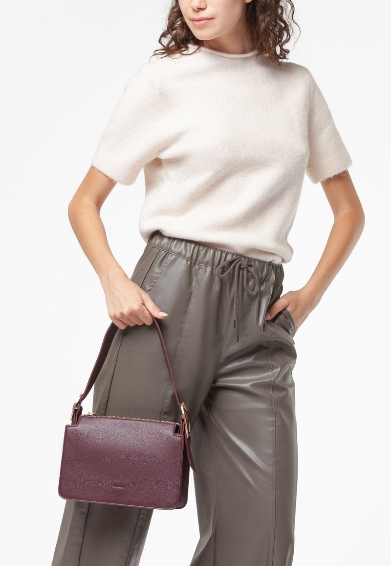 Borsa in pelle bordeaux con una forma strutturata, accessori in oro e una tracolla staccabile, tenuta da un modello che indossa un maglione chiaro e pantaloni in pelle.
