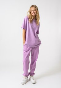 Lavandas kokvilnas t-krekls un atbilstošas bikses ar elastīgiem aprocēm; kombinētas ar baltām teksturētām sporta kedām, veidojot relaksētu, monochromatisku izskatu.