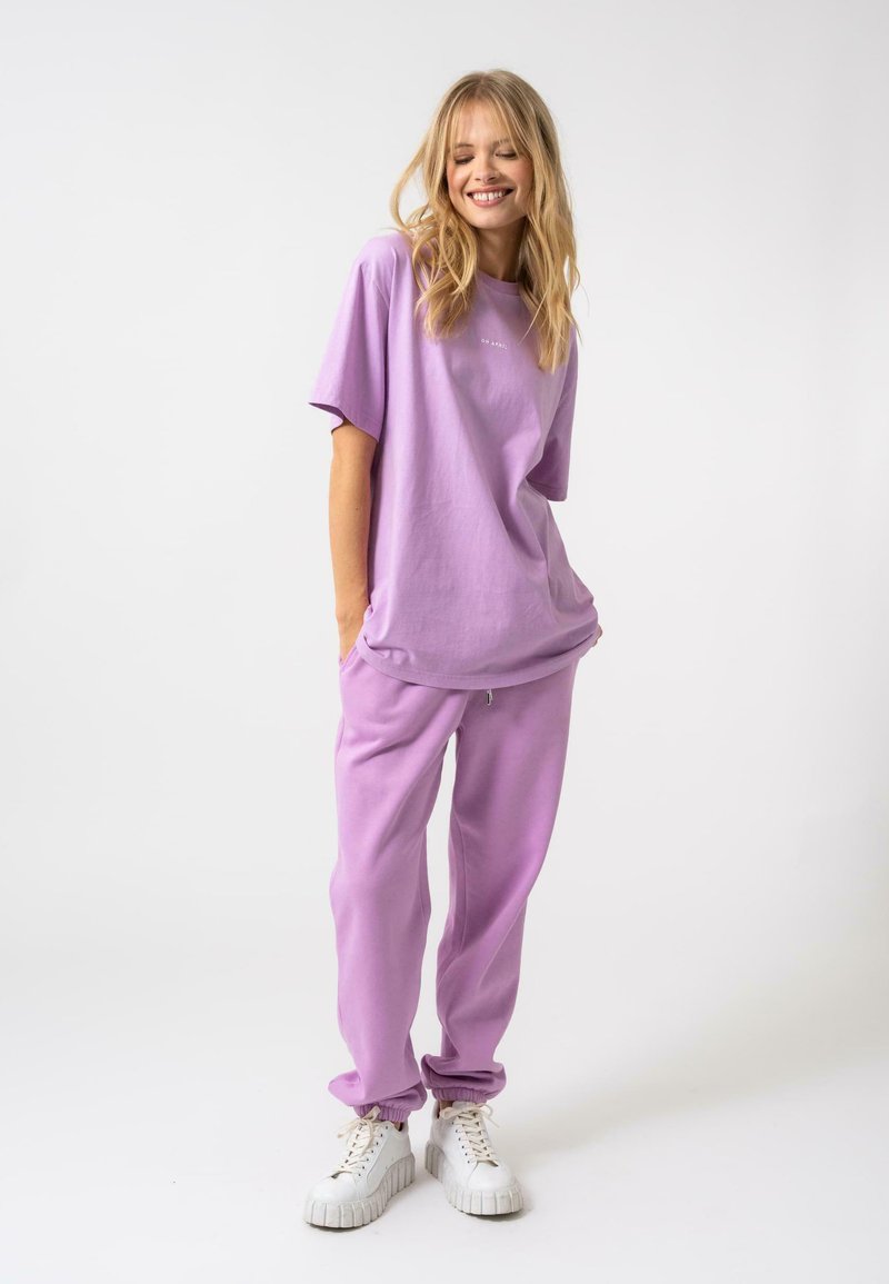 Lavandas kokvilnas t-krekls un atbilstošas bikses ar elastīgiem aprocēm; kombinētas ar baltām teksturētām sporta kedām, veidojot relaksētu, monochromatisku izskatu.