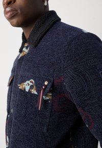 Ναυτικό μπλε μπουφάν με υφασμάτινη λεπτομέρεια, μαύρη κορυφή από σέρπα, σχέδια στις τσέπες και μετα�λλικές λεπτομέρειες.