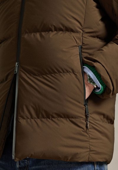 Brown puffer jacket with quilted texture, featuring a zippered pocket. Underneath, a green knit cuff is visible, paired with denim.