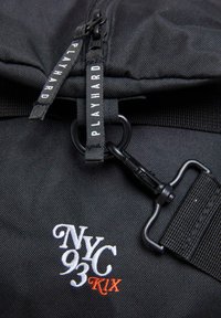 Black backpack featuring a textured fabric, dual zippers with branding, and an embroidered logo in white and orange. Includes a clip closure.
