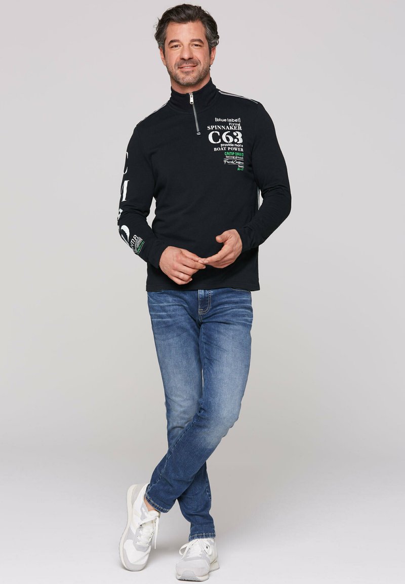 Sudadera de manga larga negra con cierre y texto impreso en blanco y verde; combinada con jeans azules y zapatillas blancas; corte relajado y textura suave.