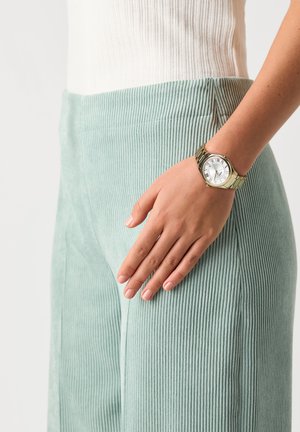 La main d'une femme reposant sur un pantalon en velours côtelé bleu clair, portant une montre-bracelet en or avec un cadran blanc et des chiffres romains.