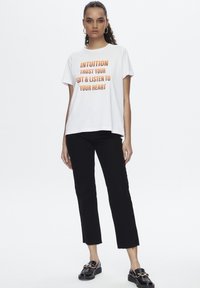 T-shirt blanche avec le texte orange "INTUITION FAIS CONFIANCE À TON INSTINCT & ÉCOUTE TON CŒUR." Associée à un pantalon noir court, des chaussures texturées et des boucles d'oreilles créoles.