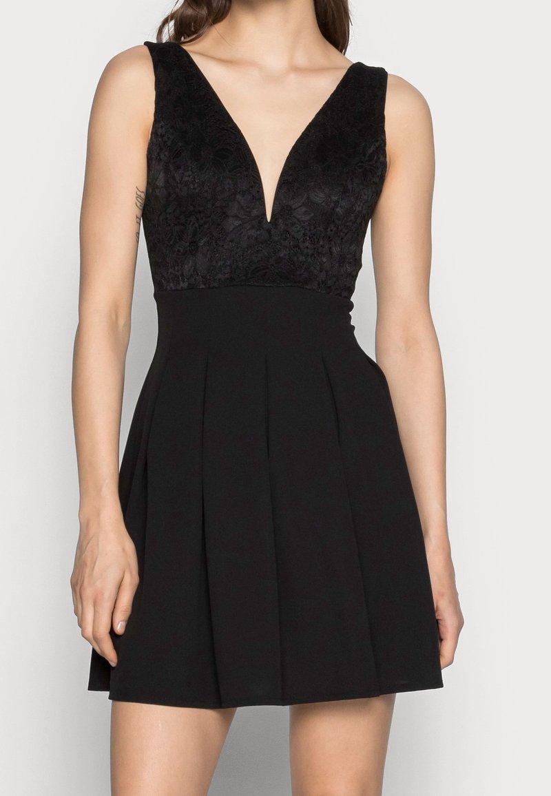 Robe noire sans manches avec corsage en dentelle à décolleté en V profond et jupe plissée portée par une femme sur un fond uni.