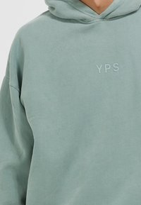 Personne portant un sweat à capuche vert clair avec les lettres en relief "YPS" sur le côté gauche de la poitrine, visible du cou au milieu du torse.