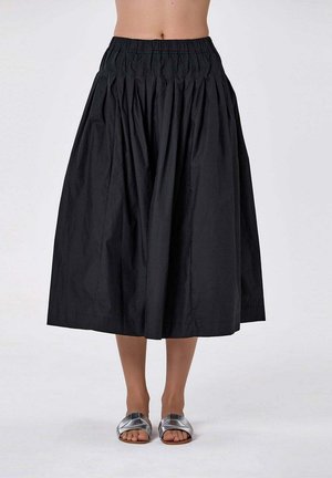 Jupe midi noire taille haute plissée portée avec des sandales plates argentées à bout ouvert sur une personne debout devant un fond uni.
