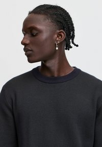 Maglione scuro a maglia con un colletto a coste, caratterizzato da un motivo testurizzato. Orecchio decorato con un orecchino a goccia di perla, modello mostrato di profilo.