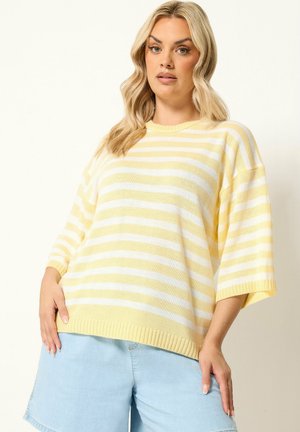 Kvinde iført gul og hvidstribet kortærmet sweater og lyseblå denimshorts, poserer med den ene hånd på hoften mod en ensfarvet baggrund.