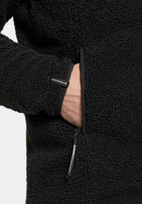 Svart fleecejacka med en texturerad yta, sidoficka och justerbar dragkedja. Har en etikett på ärmen.