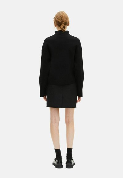 Donna con i capelli raccolti, che indossa un top nero a maniche lunghe, una gonna nera, calze nere e scarpe nere, girata di spalle su uno sfondo semplice.