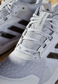 Białe buty sportowe z siateczkowymi detalami, płaskimi sznurowadłami oraz trzema czarnymi przekątnymi paskami; widoczne teksturowane materiały i wzmocnienia.