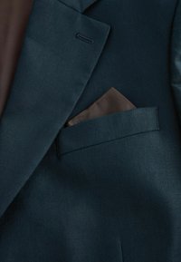 Veste de costume vert foncé avec un mouchoir de poche marron soigneusement glissé dans la poche poitrine.
