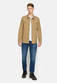 Beige skjortejakke med to brystlommer, båret over en hvid t-shirt, parret med blå jeans og brune støvler.