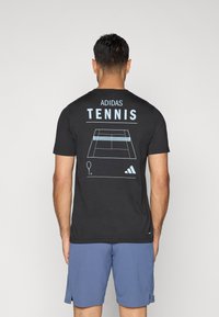 Melnā kokvilnas T-kreklā aizmugurē ir zila grafikā attēlota tenisa korta un raketes zīmējums, virs kura ir uzraksts "ADIDAS TENNIS".