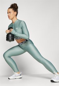 Atletische outfit in matte lichtgroene stof, bestaande uit een ritsen long-sleeve top en high-waisted leggings met een zwarte logo band. Houdt een zwarte kettlebell vast.