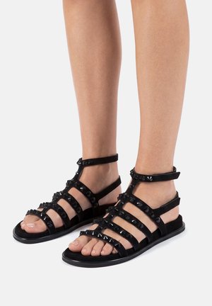 Pies con sandalias negras estilo gladiador con múltiples tiras adornadas con tachuelas cuadradas, sobre una superficie gris clara.