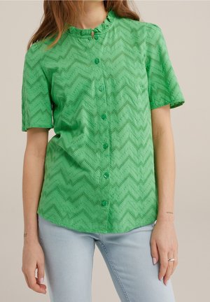 Vrouw draagt een groen zomers blouseje met korte mouwen, knopen, een zigzag-ogenpatroon en een gerimpelde kraag, gecombineerd met lichtblauwe spijkerbroek.