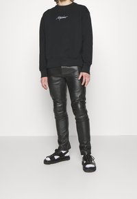 Sweat-shirt en coton noir avec logo brodé blanc, associé à un pantalon en cuir noir ajusté. Les chaussures sont des sandales noires et blanches avec des brides.