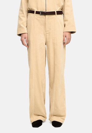 Beżowe szerokie spodnie sztruksowe w zestawie z pasującym topem, zapięte ciemnobrązowym skórzanym paskiem, noszone z czarnymi butami.