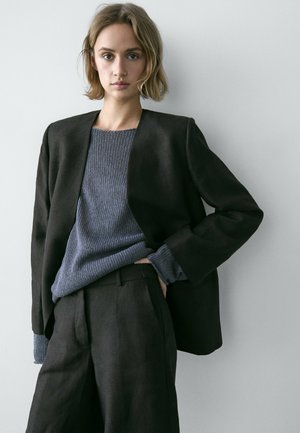 Młoda osoba z krótkimi włosami, ubrana w ciemną marynarkę na niebieskim dzianinowym swetrze i dopasowanych ciemnych spodniach, stojąca na tle jasnej ściany.