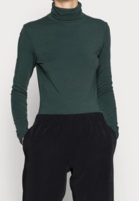 Person in einem langen, dunkelgrünen Rollkragenpullover, der in schwarze, elastische Hose gesteckt ist, mit Händen in den Taschen, vor einem schlichten Hintergrund stehend.