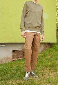Sweatshirt vert olive avec logo Nike en relief, associé à un pantalon beige clair. Le modèle porte des baskets vertes avec des lacets blancs. Fond vert.