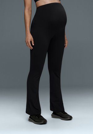 Personne enceinte portant des leggings de maternité noirs et des baskets noires, debout devant un fond sombre uni.