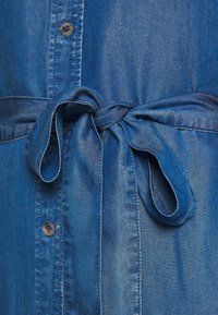 Chemise en denim bleu avec fermeture par boutons, taille ceinturée et texture douce. Dotée d'un nœud papillon à la taille et d'une légère brillance sur le tissu.