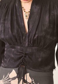 Blusa negra hecha de tela ligera con un escote en V profundo, cintura fruncida y textura sutil; presenta un lazo en la cintura.