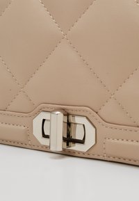 Beige quiltad läderhandväska med metallspänne och kontrasterande sömmar längs kanterna. Slät yta med en strukturerad form.